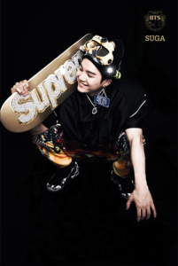 Imagem de SUGA na era BTS 2 COOL 4 SKOOL, segurando um skate dourado com o logo Supreme e usando um visual vibrante e descontraído que reflete o espírito jovem e urbano do álbum. Descubra mais no Bangtan Now.