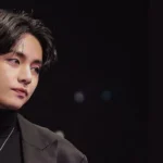 Imagem de Taehyung durante a Entrevista Taehyung SimInvest 2025, no blog do BangtanNow Bangtan Now. O artista aparece com um visual elegante em um fundo escuro, destacando sua expressão serena e presença marcante.