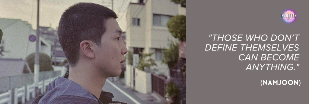 Image from Bangtan Now featuring RM (Namjoon) from BTS in a side profile, standing on a quiet residential street lined with houses and utility poles, during what appears to be early morning or late afternoon light. He wears a dark hoodie and looks contemplatively into the distance. On the right side of the image, set against a muted mauve background, there's a quote in white text: "Those who don’t define themselves can become anything." (Namjoon). The Bangtan Now logo appears in the top right corner, connecting the quote to themes of identity and introspection explored in the "RM: Right People Wrong Person" documentary.