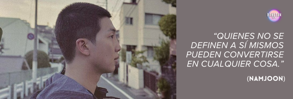 Imagen de Bangtan Now con RM (Namjoon), de BTS, de perfil, parado en una calle residencial tranquila, rodeada de casas y postes eléctricos, bajo la luz suave del amanecer o del atardecer. Lleva una sudadera oscura con capucha y mira contemplativamente a lo lejos. En el lado derecho de la imagen, sobre un fondo malva apagado, aparece una cita en texto blanco: “Quienes no se definen a sí mismos pueden convertirse en cualquier cosa.” (Namjoon). El logo de Bangtan Now está en la esquina superior derecha, conectando la frase con los temas de identidad e introspección explorados en el documental RM: Right People Wrong Person.