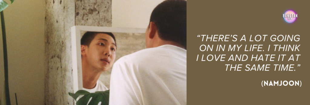 Image from Bangtan Now showing RM (Namjoon) from BTS in an introspective moment. He is seen from behind, gazing into a small mirror propped against a concrete wall, where his reflection looks thoughtfully back. He wears a plain white T-shirt, and a large plant leaf partially frames the bottom of the mirror. The scene is softly lit, evoking a sense of quiet self-reflection. On the right, against a brown background, appears a quote in white italic text: “There’s a lot going on in my life. I think I love and hate it at the same time.” (Namjoon). The Bangtan Now logo sits in the upper corner, tying the image to the "RM: Right People Wrong Person" documentary.