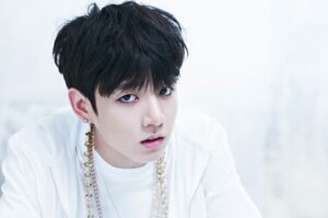 Jungkook, integrante do BTS, em um ensaio promocional para o bts o rul82 album, com figurino branco e acessórios dourados, representando o conceito icônico da era. Imagem destacada no site Bangtan Now.