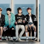 Imagem de capa do artigo sobre Spring Day, do BTS, publicada pelo Bangtan Now. A foto mostra os sete integrantes do grupo sentados em uma estrutura aberta, com o mar ao fundo, transmitindo uma sensação de nostalgia e amizade. O design inclui um fundo azul suave com o título "Álbum You Never Walk Alone – Spring Day", destacando a conexão da música com o álbum e seu impacto emocional.