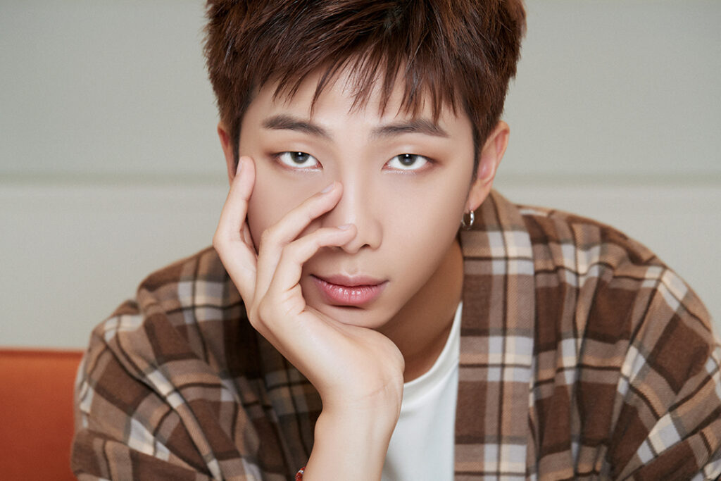 Agradecimentos álbum BE - Bangtan Now (2) Imagem de RM, líder do BTS, com uma expressão serena e olhar penetrante. Ele veste uma camisa xadrez em tons terrosos sobre uma camiseta branca e apoia a mão no rosto, criando uma pose introspectiva. A atmosfera suave da foto reflete a profundidade emocional presente nos agradecimentos de RM no álbum BE BTS, onde ele compartilhou suas reflexões sobre a pandemia e a importância da conexão com o ARMY. Essa imagem faz parte do artigo sobre os agradecimentos BE BTS, destacando as mensagens de gratidão e esperança expressas pelos membros durante a criação do álbum. Imagem ilustrativa do artigo do Bangtan Now.