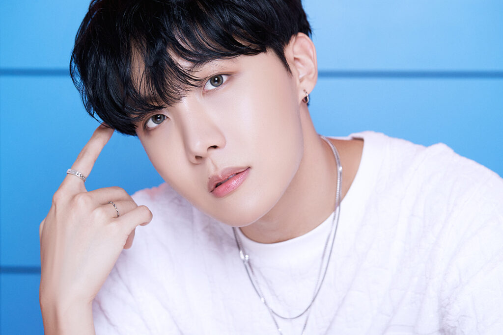 Agradecimentos álbum BE - Bangtan Now (5) Imagem de J-Hope, integrante do BTS, com uma expressão serena e olhar cativante. Ele veste uma camiseta branca e acessórios prateados, incluindo colares e anéis, enquanto toca levemente a lateral da cabeça com um dos dedos. O fundo azul vibrante cria um contraste harmonioso com seu visual. Essa foto faz parte do artigo sobre os agradecimentos de J-Hope no álbum BE BTS, onde ele compartilha palavras de encorajamento e gratidão ao ARMY, destacando a importância de seguir em frente com esperança. No contexto dos agradecimentos BE BTS, J-Hope reforça seu desejo de levar conforto através da música e expressa sua imensa gratidão por todos que fizeram parte desse projeto. Imagem que ilustra o artigo do Bangtan Now.