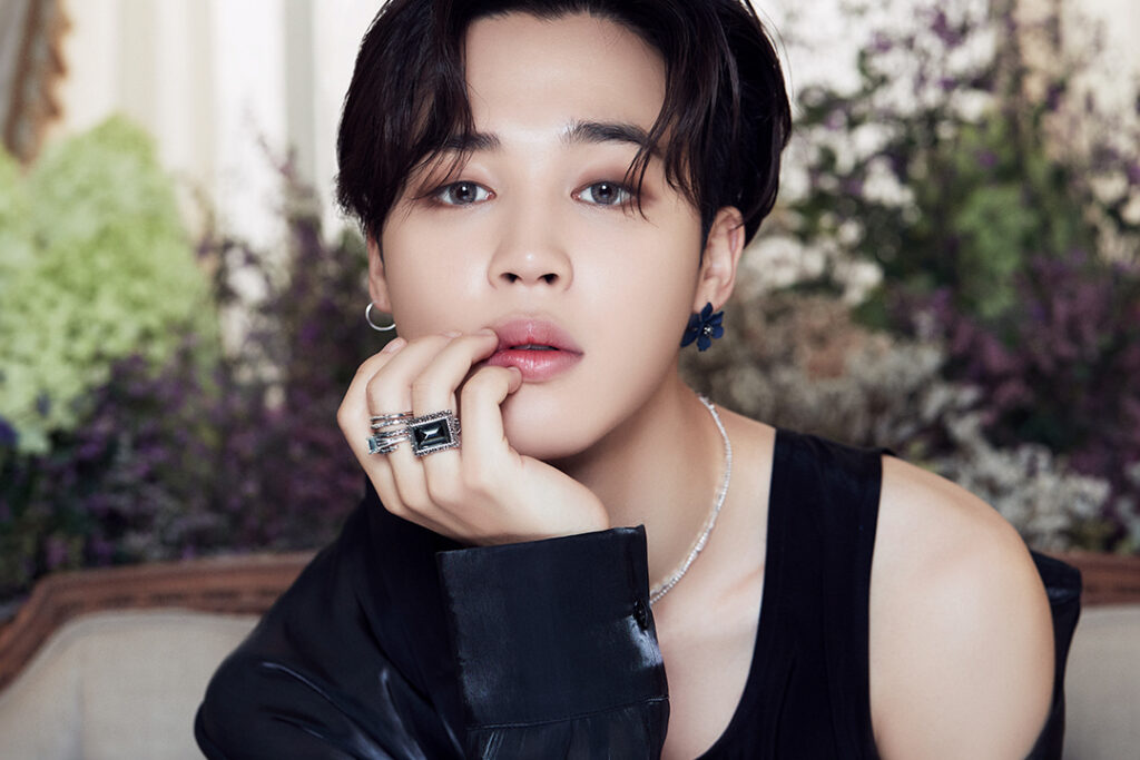 Agradecimentos álbum BE - Bangtan Now (6) Imagem de Jimin, integrante do BTS, com uma expressão delicada e olhar profundo. Ele veste uma regata preta com uma jaqueta de couro caída sobre um dos ombros, além de acessórios sofisticados como anéis prateados, colar brilhante e brincos florais. Sua mão repousa suavemente no rosto, enfatizando a atmosfera elegante e contemplativa da imagem. O fundo é composto por flores e elementos naturais, criando um contraste entre o visual moderno e a estética suave da cena. Essa foto faz parte do artigo sobre os agradecimentos de Jimin no álbum BE BTS, onde ele expressa sua gratidão e carinho ao ARMY, destacando a importância do apoio mútuo. No contexto dos agradecimentos BE BTS, Jimin compartilha sua emoção e desejo de continuar crescendo ao lado dos fãs. Imagem que ilustra o artigo do Bangtan Now.