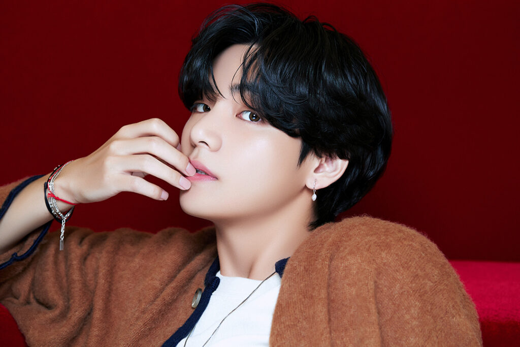 Agradecimentos álbum BE - Bangtan Now (7) Imagem de V (Taehyung), integrante do BTS, com um olhar intenso e expressão serena. Ele veste um cardigã marrom sobre uma camiseta branca e usa acessórios delicados, incluindo um brinco de pérola e pulseiras. Sua pose descontraída, com a mão próxima aos lábios, contrasta com o fundo vermelho vibrante, criando uma estética sofisticada e artística. Essa foto faz parte do artigo sobre os agradecimentos de Taehyung no álbum BE BTS, onde ele compartilha seu amor pelo ARMY e a saudade dos palcos. No contexto dos agradecimentos BE BTS, V expressa sua esperança e a promessa de reencontro com os fãs. Imagem que ilustra o artigo do Bangtan Now.