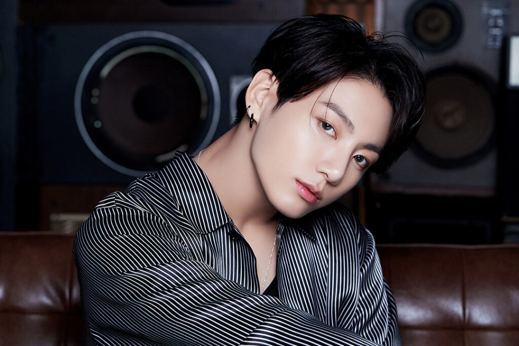 Agradecimentos álbum BE - Bangtan Now (8) Imagem de Jungkook (JK), o maknae do BTS, com um olhar expressivo e postura relaxada. Ele veste uma camisa listrada preta e branca, levemente aberta, e acessórios minimalistas como um brinco preto e um colar prateado. Seu cabelo está penteado para trás, destacando seu rosto iluminado. Ao fundo, grandes caixas de som reforçam a estética musical da cena. Essa foto faz parte do artigo sobre os agradecimentos de Jungkook no álbum BE BTS, onde ele compartilha sua imensa gratidão ao ARMY e sua vontade de se manter conectado por meio da música. No contexto dos agradecimentos BE BTS, JK expressa sua saudade dos fãs e o desejo de reencontrá-los em breve. Imagem que ilustra o artigo do Bangtan Now.