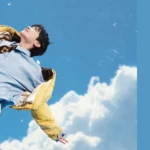 Capa do artigo sobre Sweet Dreams, colaboração de J-Hope e Miguel, publicada pela Bangtan Now. A imagem mostra J-Hope flutuando no céu, com os braços abertos e uma expressão serena, refletindo a estética sonhadora do MV. O fundo azul com nuvens remete ao conceito visual da música, transmitindo leveza e liberdade.