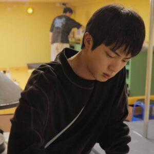 Jin aparece concentrado en una tarea dentro de la posada de Kian’s Bizarre B&B, escenario del reality Jin BTS Netflix. Viste un buzo oscuro y se encuentra en un ambiente interior iluminado por una luz amarilla. Al fondo, otro participante está cocinando. La imagen, compartida por el blog Bangtan Now, muestra uno de los momentos más tranquilos y reflexivos del programa.