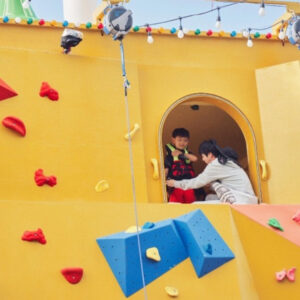 Escena de Kian’s Bizarre B&B, con un niño equipado para escalar de pie en la entrada de una colorida posada amarilla con una pared de escalada. Jin lo ayuda con cuidado y atención. El escenario lúdico, parte del reality show de Netflix, resalta los momentos cálidos y entretenidos que definen a Jin BTS Netflix, en una imagen destacada por el blog Bangtan Now.