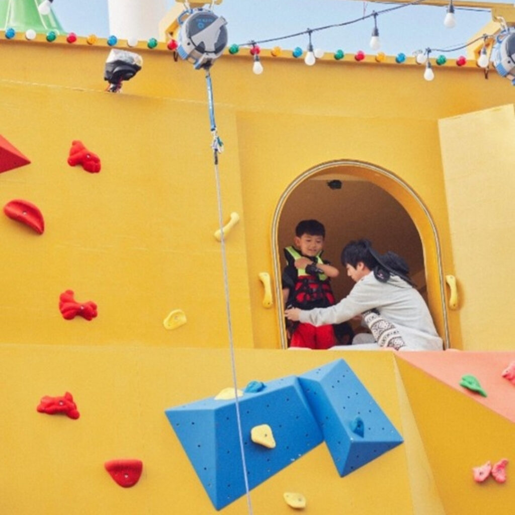 Jin BTS Netflix - Foto 3 - Bangtan Now Jin ajuda uma criança a se preparar para descer por uma parede de escalada colorida, na entrada da pousada do reality Kian’s Bizarre B&B. A estrutura amarela, com apoios vermelhos, azuis e amarelos, compõe o cenário do programa Jin BTS Netflix, divulgado pelo Bangtan Now.