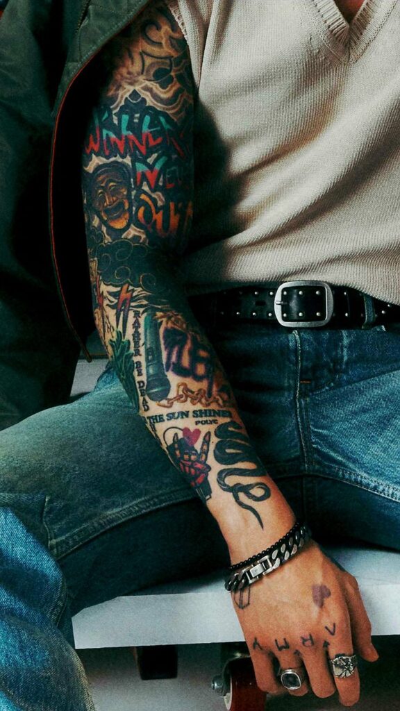 BTS Tatoos - Bangtan Now (1) Close-up of Jungkook’s fully tattooed right arm and hand, seated and dressed in a beige knit shirt and blue jeans. The tattoo sleeve features a mix of bold, colorful designs, including graffiti-style text, a mask, lightning bolt, flames, and meaningful phrases like “Rather be dead than cool” and “The sun shines.” His fingers display the word “ARMY” and a small purple heart. This detailed shot is from the “Golden” album photoshoot and is featured in the Bangtan Now article on BTS Tattoos, highlighting Jungkook’s evolving tattoo journey.