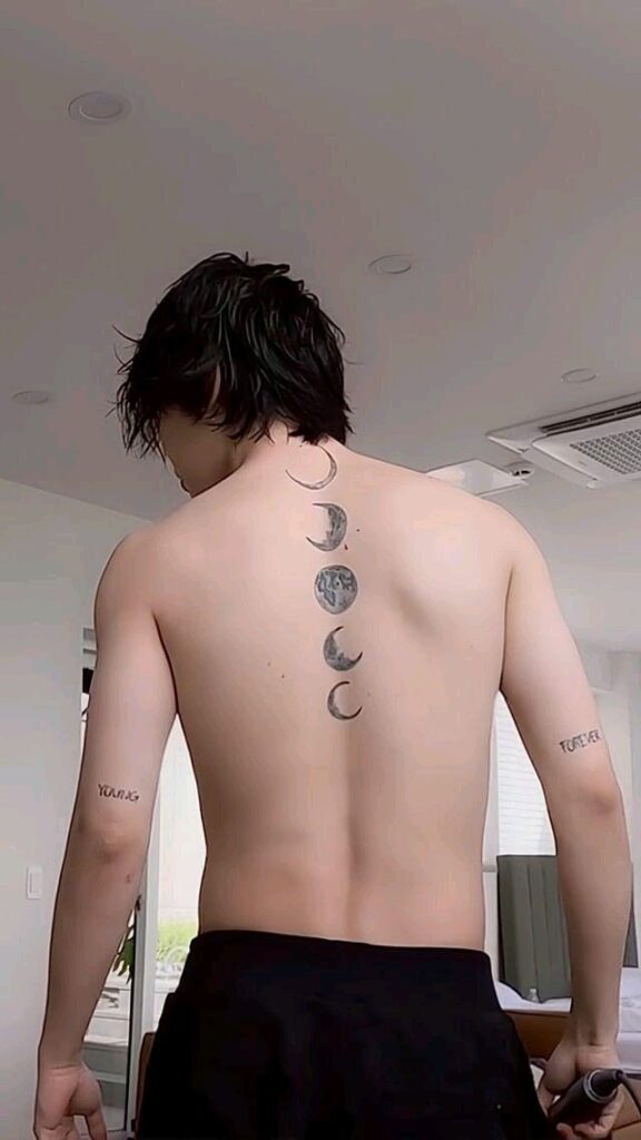 BTS Tatoos - Bangtan Now (3) Back view of BTS member Jimin standing shirtless in a bright room, revealing a tattoo of five moon phases vertically aligned along his spine — from crescent to full and back. The words “YOUNG” and “FOREVER” are also tattooed on the back of his arms. This scene is from the reality show “Are You Sure” with Jungkook and V, and is featured in a Bangtan Now article exploring the personal stories behind BTS Tattoos.