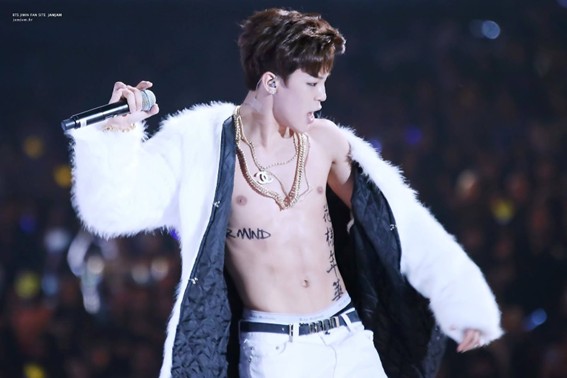 BTS Tatoos - Bangtan Now (4) Jimin of BTS performing shirtless at the 2014 MAMA Awards, wearing a white fur coat and layered gold chains, while revealing a large temporary tattoo that reads “NEVERMIND” across his ribs. This early moment became iconic among fans, marking one of his first public expressions through body art. The image appears in a Bangtan Now article on BTS Tattoos, showcasing the evolution of each member’s relationship with tattoos.