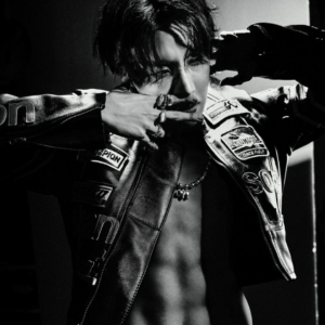 Close-up black-and-white concept photo of j-hope for Killin it Girl music, spotlighted in the Bangtan Now article. Wearing a leather racing jacket open to reveal his torso, he poses with both hands near his face, adorned with chunky rings. The dramatic lighting enhances the photo’s bold, edgy tone, capturing the essence of confidence and allure in this era.