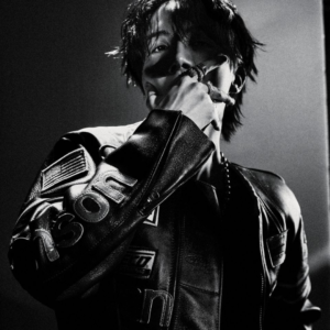 j-hope en una concept photo en blanco y negro de la música Killin it Girl, publicada en el artículo de Bangtan Now. Luce una chaqueta de cuero con parches y posa con la mano cerca del rostro, mostrando anillos llamativos. La iluminación dramática y el encuadre refuerzan el aire rebelde y sofisticado de esta etapa visual.