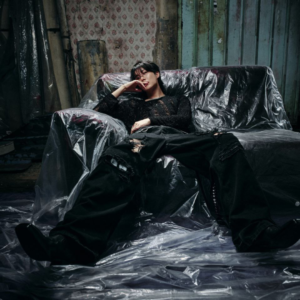 j-hope en una concept photo de la música Killin it Girl, compartida en el artículo de Bangtan Now. Reclinado en un sofá cubierto con plástico, en un ambiente rústico y descuidado, viste un top brillante y pantalones oscuros rotos. La escena transmite una mezcla de vulnerabilidad, poder y rebeldía visual, alineada con el tono provocador y artístico de esta era.