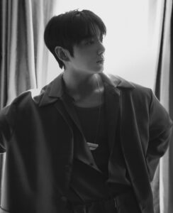 Black and white photograph of Jungkook Calvin Klein backstage at the brand’s 2025 fashion show, published on the Bangtan Now blog. He is shown in profile, standing in front of a curtained window with soft natural light illuminating his face in a gentle, dramatic way. Jungkook wears a dark tailored outfit with a modern-cut shirt and blazer over a tank top. He accessorizes with layered necklaces, ear piercings, and a visible lip piercing, enhancing his elegant, contemporary look. His expression is calm and contemplative, capturing a quiet moment of reflection before the event.