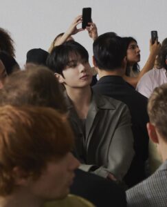 Color photograph of Jungkook Calvin Klein watching the brand’s 2025 fashion show, featured on the Bangtan Now blog. Seated among the audience, he wears a sophisticated gray suit and has his arms crossed, gazing attentively toward the runway out of frame. His hair is neatly styled to the side, with visible silver hoop earrings and lip piercings. In the background, several attendees are holding up phones to capture the moment. The image reflects the elegant and exclusive atmosphere of the event.