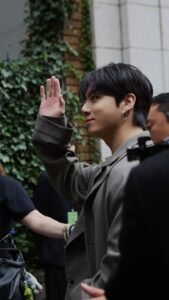 Color photograph of Jungkook Calvin Klein arriving at the brand’s 2025 fashion show, published on the Bangtan Now blog. He is shown in profile, smiling gently and waving with his right hand. He wears a modern-cut gray suit, with his dark hair styled to the side, silver hoop earrings, and visible lip piercings. In the background, there’s a brick wall covered in greenery and members of the production team. The image captures a warm, welcoming moment of grace and approachability.