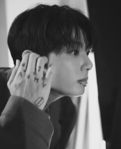 Black and white photograph of Jungkook Calvin Klein behind the scenes at the brand’s 2025 fashion show, published on the Bangtan Now blog. He is shown in profile, gazing intently at something outside the frame with a focused expression. His hand is raised near his face, displaying tattoos on his fingers, including the letters “A.R.M.Y.” and a geometric design. He wears a dark tailored suit, and a lip piercing is visible. Soft lighting highlights his facial features, creating an intimate and contemplative mood.