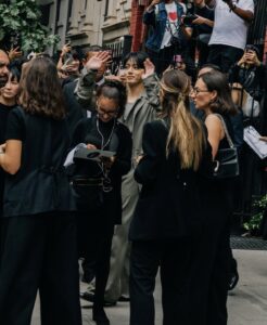 Color photograph of Jungkook Calvin Klein arriving at the brand’s 2025 fashion show, surrounded by fans and production staff. He is wearing an elegant light gray tailored suit and waving with both hands, smiling as he walks through the crowd. The atmosphere is bustling, with people taking photos, filming, and holding event programs. The image captures the excitement surrounding his presence. Photo published on the Bangtan Now blog.