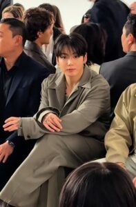Color photograph of Jungkook Calvin Klein seated in the front row of the brand’s 2025 fashion show. He is wearing an elegant light gray tailored suit, sitting with his legs crossed and looking directly at the camera with a calm and attentive expression. The sleeves of his blazer are slightly rolled up, revealing part of the tattoos on his hands. Around him, other guests are also watching the show, reinforcing the sophisticated atmosphere of the event. Image published on the Bangtan Now blog.