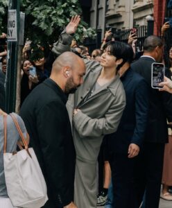 Color photograph of Jungkook Calvin Klein waving to fans outside the venue of the brand’s 2025 fashion show, published on the Bangtan Now blog. He is wearing a light khaki-colored tailored suit and smiling subtly while raising his right hand in greeting. Jungkook is surrounded by security guards and a large crowd of enthusiastic fans, many holding up their phones to capture the moment. The image conveys the vibrant energy of his arrival, highlighting his charismatic presence and the refined style that defined his appearance at the event.