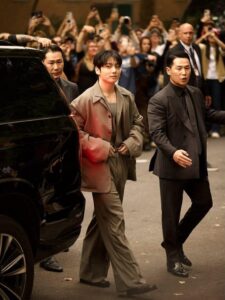 Color photograph of Jungkook Calvin Klein arriving at the brand’s 2025 fashion show, featured on the Bangtan Now blog. He is walking beside his manager, Mr. Lee, surrounded by security guards, while a large crowd of fans in the background captures the moment with their phones. Jungkook wears a modern, elegant brown tailored suit with a loose blazer, straight-cut trousers, and a matching tank top underneath. His expression is calm and confident, with visible lip piercings and perfectly styled hair. The scene is bathed in warm lighting, emphasizing his sophisticated look amid the buzz of the event.