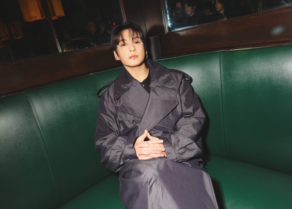 Jungkook Calvin Klein 2025 - Bangtan Now (8) Color photograph of Jungkook Calvin Klein during the after-party of the brand’s 2025 fashion show. He is seated on a green leather sofa with his hands folded on his lap, wearing an elegant black overcoat. The lighting highlights his calm face and direct gaze into the camera. In the background, a mirror reflection reveals other guests in the room, creating a sophisticated and intimate atmosphere. Photo published on the Bangtan Now blog.