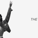 Black and white promotional image showing Jungkook from BTS, shirtless and wearing an unbuttoned Calvin Klein denim jacket and jeans. He stands confidently with both arms raised high in a V-shape, looking straight ahead with a calm, assertive expression. His toned abs are visible, and the Calvin Klein logo can be seen on the waistband of his underwear and on the jacket pocket. To the right, the image features the article title in bold text: “Jungkook Calvin Klein – The Partnership That Took the Fashion World by Storm,” along with the Bangtan Now logo, signaling the article’s publication source.