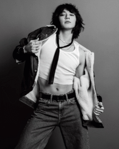 Black and white photo of Jungkook Calvin Klein from the brand’s campaign, posing front-facing with a firm expression and direct gaze at the camera. He is wearing an open denim jacket with a shearling lining, revealing a fitted white tank top and a thin black tie hanging loosely around his neck. His dark-wash jeans are paired with a black belt. Multiple rings adorn his fingers, and his tattoos are subtly visible. The neutral gray background highlights the contrast in lighting and the artist’s striking look. Image published on the Bangtan Now blog.