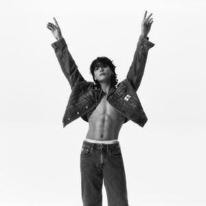 Black and white photo of Jungkook Calvin Klein from the brand’s campaign, posing with his arms raised and fingers pointing upward, in front of a white background. He is wearing an open Calvin Klein denim jacket, revealing his defined abs, and low-rise jeans that expose the waistband of matching Calvin Klein underwear. His hair is loose, with bangs falling over his forehead, and his expression is calm as he looks slightly upward. Image published on the Bangtan Now blog.