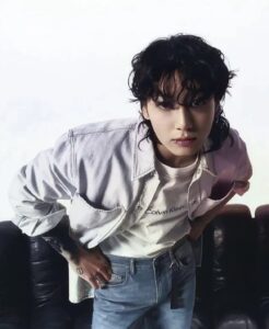 Color photo of Jungkook Calvin Klein from the brand’s campaign, leaning forward with his hands resting on his waist. He is wearing a white T-shirt with the “Calvin Klein” logo centered on the chest, a light denim shirt open over it, and blue jeans. His hair is loose and wavy, with bangs falling over his forehead. Visible tattoos can be seen on his right hand, and he gazes directly at the camera with a focused expression. In the background, there is a black sofa against a softly blurred white backdrop. Image published on the Bangtan Now blog.