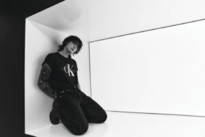 Black and white photo of Jungkook Calvin Klein from the brand’s campaign, kneeling inside a softly lit white cube structure. He is wearing a black T-shirt with a large “CK Calvin Klein Jeans” logo in the center, straight-cut black pants, and chunky-soled boots. His fully tattooed right arm is visible, resting behind his body. His hair is loose and slightly wavy, and he gazes forward with a calm expression. Image published on the Bangtan Now blog.