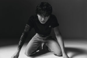 Black and white image of Jungkook Calvin Klein from the brand’s campaign, kneeling on the floor of a studio with dramatic lighting. He is wearing a black T-shirt with the “CK” logo on the left side of the chest and light-wash jeans. His hair is wet and messy, with bangs falling over his eyes, and he stares intensely at the camera. The full tattoo sleeve on his right arm is completely visible, showcasing intricate designs. The pose conveys strength and presence. Image published on the Bangtan Now blog.