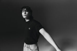 Black and white photograph of Jungkook Calvin Klein posing with a calm expression in a studio with a dark background. He is wearing a black T-shirt with the embroidered “CK” logo on the chest and high-waisted light-wash jeans. His left arm is extended out of frame, creating a subtle sense of movement. The lighting highlights his features and the contrast between the elements of his minimalist look. Image published on the Bangtan Now blog.