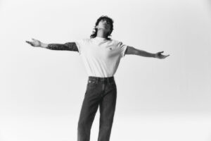 Black and white photo of Jungkook Calvin Klein from the brand’s campaign, with arms open in an expansive pose and his face slightly tilted upward. He is wearing a white T-shirt with the “CK” logo on the left side of the chest and dark jeans. His fully tattooed right arm is prominently displayed, standing out against the minimalist white background. The pose conveys a sense of freedom and confidence. Image published on the Bangtan Now blog.