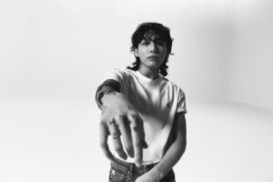 Black and white photo of Jungkook Calvin Klein from the brand’s campaign, extending his arm toward the camera with a focus on the tattoos on his hands and fingers, including the letters “ARMY.” He is wearing a white T-shirt with the “CK” logo on the chest and mid-rise jeans, with the waistband of his Calvin Klein underwear slightly visible. The background is white and minimalist, emphasizing his presence and calm expression. Image published on the Bangtan Now blog.