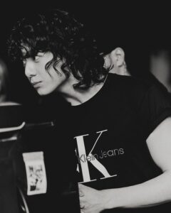 Black and white behind-the-scenes photo of Jungkook Calvin Klein from the brand’s campaign, wearing a T-shirt with the “Klein Jeans” logo prominently displayed. He is slightly leaning forward, with a focused gaze directed at something off-camera. His hair is loose and wavy, falling over his face. The lighting is soft, and the background is dark and blurred, suggesting a studio setting. Image published on the Bangtan Now blog.