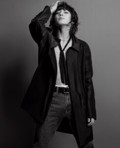 Black and white photo of Jungkook Calvin Klein, posing with a confident expression and his gaze directed upward. One hand rests on his wavy hair, and he wears an open black overcoat with no shirt underneath, revealing part of his chest and a thin black tie hanging loosely. His high-waisted dark jeans are paired with a black belt featuring a metallic buckle. Soft lighting highlights the fabric textures and the contours of his face. Image published on the Bangtan Now blog.