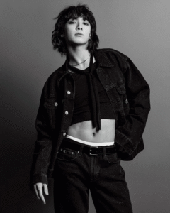 Black and white photo of Jungkook Calvin Klein from the brand’s campaign, posing casually against a plain gray background. He is wearing a dark denim set with an open jacket, a black cropped top with a neck tie, and mid-rise jeans that reveal the waistband of his Calvin Klein underwear. His hair is loose, wavy, and voluminous. He wears a necklace, a hoop earring, and looks confidently into the camera. Image published on the Bangtan Now blog.