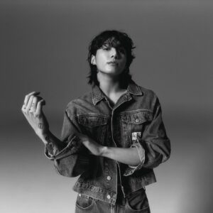 Jungkook Calvin Klein posa em um estúdio com fundo cinza, em foto em preto e branco da campanha da marca. Ele veste um conjunto jeans da Calvin Klein com jaqueta estruturada, parcialmente aberta, e calça de cintura média. Seu cabelo está solto e levemente bagunçado, com franja cobrindo parte da testa. Jungkook encara a câmera com expressão séria e segura a manga da jaqueta com uma das mãos, exibindo tatuagens visíveis nos dedos e antebraço. Imagem publicada no blog Bangtan Now.