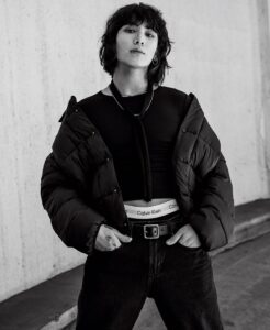Black and white photo of Jungkook Calvin Klein from the brand’s campaign, posing with attitude in front of a concrete wall. He is wearing a fitted black cropped top with a neck tie, a voluminous puffer jacket draped over his shoulders, dark jeans, and a belt with a buckle. The waistband of his Calvin Klein underwear is visible above the jeans. His hair is loose and wavy, with bangs partially covering his forehead. He wears a lip piercing, a hoop earring, and a necklace. Image published on the Bangtan Now blog.
