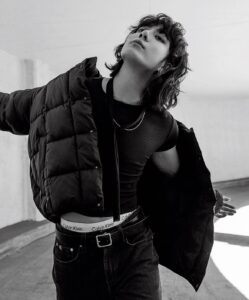 Black and white photo of Jungkook Calvin Klein, wearing dark jeans, a fitted black T-shirt, and a puffer jacket draped over one shoulder. His white Calvin Klein underwear is intentionally visible above the waistband of his jeans. He poses with arms open and his face tilted upward, displaying a striking gaze. The setting is an outdoor concrete passageway lit by natural light. Image published on the Bangtan Now blog.