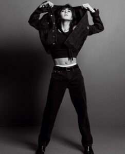 Black and white photo of Jungkook Calvin Klein from the brand’s campaign, taken in a studio with a gray background. He stands with his legs apart, lifting a dark denim jacket to reveal a fitted black cropped top and the waistband of his Calvin Klein underwear showing above his jeans. The look combines attitude and style, emphasizing the contrast between the tight-fitting top and the straight-cut jeans. His gaze is directed upward, and the pose conveys confidence and presence. Image published on the Bangtan Now blog.