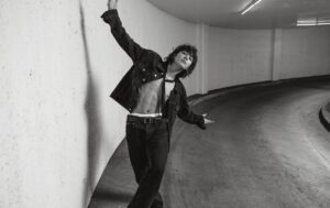 Black and white photo of Jungkook Calvin Klein from the brand’s campaign, posing in a curved garage area under artificial lighting. He is wearing a dark denim set with an open jacket, revealing his defined abs and the waistband of his Calvin Klein underwear. He has on a black cropped top with ties and a belt with a buckle. With arms open wide, Jungkook leans back slightly against the wall in an expressive, artistic pose. Image published on the Bangtan Now blog.