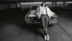 Black and white photo of Jungkook Calvin Klein from the brand’s campaign, leaning against a classic car parked in a dark garage. He is wearing a white T-shirt with the “CK” logo on the chest, an open light-colored puffer jacket, straight-cut jeans, a black belt, and white sneakers. His body is slightly leaned back with his arms resting on the hood, and his head tilted upward with eyes closed, conveying a sense of relaxation and attitude. Image published on the Bangtan Now blog.