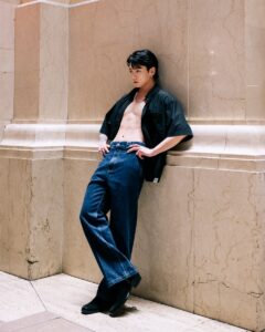 Color photo of Jungkook Calvin Klein from the brand’s campaign, leaning against a beige marble wall with a confident expression. He is wearing an open black shirt that reveals his defined abs, straight-cut dark-wash jeans, and black boots. The tattoos on his right arm are partially visible, and his hair is slicked back for a more polished look. Soft lighting highlights the contrast between his modern outfit and the classic setting. Image published on the Bangtan Now blog.